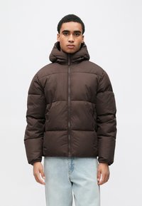 Καφέ μπουφάν τύπου puffer με κουκούλα, με τετράγωνο σχήμα, καπιτονέ σχέδιο, εμπρόσθιο φερμουάρ και ριμπ μανσέτες. Φοριέται πάνω από ανοιχτόχρωμα μπλε τζιν.