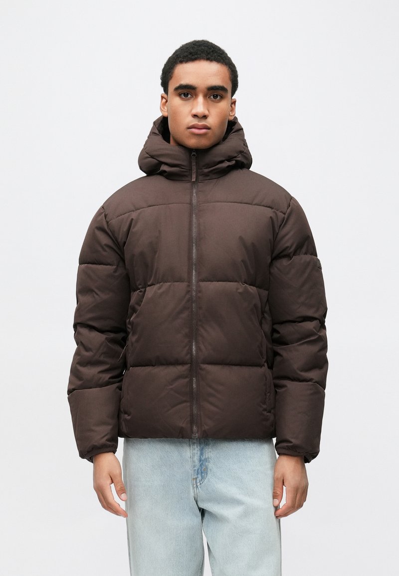 Καφέ μπουφάν τύπου puffer με κουκούλα, με τετράγωνο σχήμα, καπιτονέ σχέδιο, εμπρόσθιο φερμουάρ και ριμπ μανσέτες. Φοριέται πάνω από ανοιχτόχρωμα μπλε τζιν.