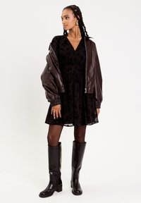 Svart blommig klänning med en utsvängd kjol, ihop med en brun läderbomberjacka och knähöga svarta stövlar. Enhetlig monokrom look.