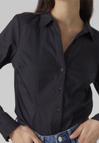 Camicia nera con colletto, caratterizzata da maniche lunghe, texture liscia e cuciture sottili. Abbinata a jeans blu a vita alta.