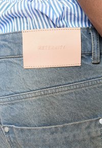 Denimjeans med en lys blå vask har en pink læderlogo-patch mærket "RETERNITY" på bagsiden af taljen. Stikningerne er synlige.