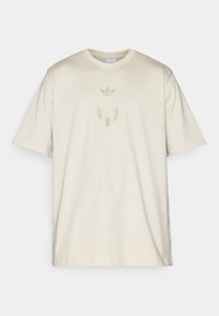 Beige puuvillane T-särk, millel on ümmargune kaelus, esiküljel on tõstetud Adidas logo ja embleem, mis on trükitud sobivas värvitoonis.
