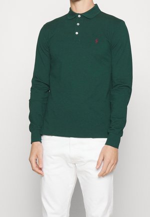 Camisa polo de manga larga de color verde oscuro con cuello, que presenta un pequeño logotipo rojo. Combinada con pantalones blancos. Está hecha de una tela suave.