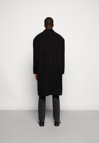 Schwarzer langer Mantel mit hohem Kragen, aus strukturiertem Stoff. Trägt eine graue Hose und schwarze Schuhe, von hinten betrachtet.