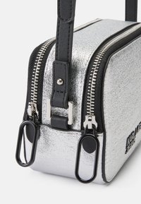 Sølvmetallic veske med rektangulær form, svarte skinnaccenter, doble glidelåser og stilige metalldetaljer. Glatt tekstur overalt.