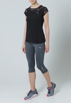 Maglietta atletica nera con dettagli in rete trasparente sulle spalle, abbinata a leggings grigi fino al ginocchio con accenti rosa e un logo. Scarpe da ginnastica grigie con suole rosse.