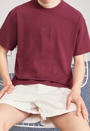 T-shirt en coton couleur bordeaux avec logo Nike brodé, coupe décontractée, assorti à un short beige clair. Sujet assis, entouré de meubles de couleur claire.