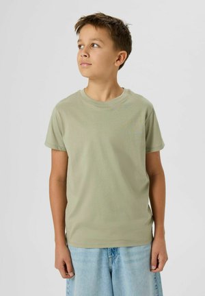 Fant z kratkimi rjavimi lasmi, oblečen v svetlo bež majico in svetle modre kavbojke, ki rahlo gleda navzgor in levo, na enobarvni podlagi.