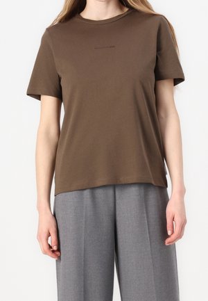 Kobieta ubrana w brązowy t-shirt z krótkim rękawem i szare szerokie spodnie, stojąca na jednolitym białym tle.