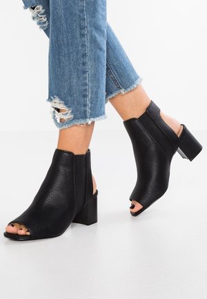 Sorte peep-toe ankelstøvler lavet af tekstureret læder med blokhæl, der har åbne sider og en glat finish. Kombineret med slidte blå jeans.
