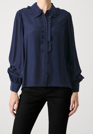 Laivastonsininen pusero, jossa on röyhelökaulus ja napillinen etulista, pitkät puhvimaiset hihat, joissa on kerätyt kalvosimet, yhdistettynä mustiin housuihin.