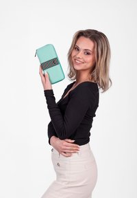 Young woman with blonde hair smiling, holding a turquoise wallet with a colorful zigzag pattern against a plain white background.