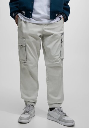 Personne portant un pantalon cargo gris clair avec des poches latérales, des baskets blanches et grises, et une veste sombre, les mains dans les poches.