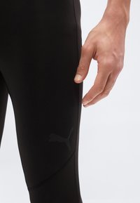 Κοντινό πλάνο ενός χεριού δίπλα σε μαύρο αθλητικό κολάν με διακριτικό λογότυπο Puma στον μηρό, πάνω σε απλό φόντο.