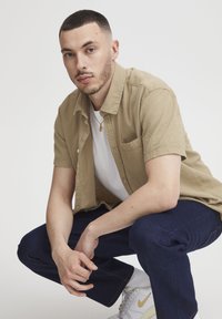 Man met kort haar en neusring die in squat positie zit, gekleed in een beige overhemd met korte mouwen, een witte t-shirt, donkere jeans en witte sneakers met een gele swoosh.
