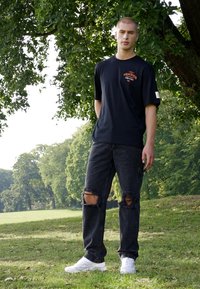 Black cotton t-shirt with printed red text, paired with ripped black jeans and white sneakers, set against a green park backdrop.