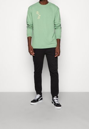 Vihreä pitkähihainen paita, jossa tekstiprintti, yhdistetty mustiin cargo-housuihin ja mustiin korkeavartisiin lenkkareihin, joissa on valkoiset nauhat ja pohjat.