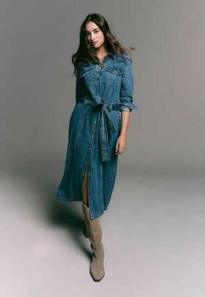 Rochie din denim cu mâneci lungi, guler cu nasturi, talie evidențiată cu curea și fente laterale. Purtați cu cizme bej din suede, până la genunchi.
