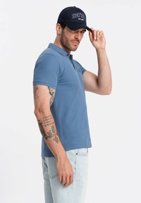 Modrá polokošile s klasickým límečkem a vyšitým logem. K tomu je nošena námořnická čepice s nápisem "NEW JERSEY" a zakřiveným kšiltem.