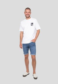 T-shirt blanche à manches courtes avec un motif imprimé sur la poche, associé à un short en denim retroussé et des chaussures légères à enfiler. Fond neutre.