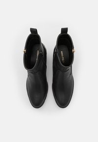Botas de tobillo de cuero negro con textura suave, cremalleras laterales y pequeñas lengüetas en los tobillos. Acentos dorados en las cremalleras.