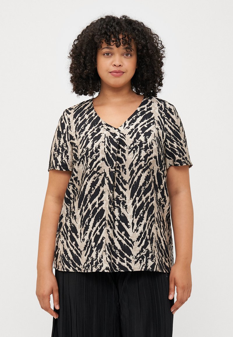 Une femme aux cheveux bouclés porte un haut à manches courtes et décolleté en V, arborant un imprimé animal audacieux noir et beige, associé à un pantalon plissé noir.
