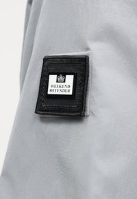 Μαύρο και άσπρο μπαλώμα "Weekend Offender" με έμβλημα κορώνας ραμμένο σε ανοιχτό γκρι ύφασμα, πιθανώς μανίκι μπουφάν.