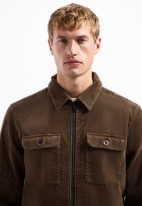 Bruine corduroyjas met een rits aan de voorkant, twee borstzakken met knoopsluitingen en een kraag. Gelaagd stof en verticale ribbel.
