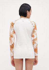 Dug dug rukav rash guard u krem boji s narančastim cvjetnim uzorkom na rukavima. Ima okrugli izrez i gladak, rastezljiv materijal.