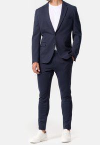 Blazer bleu marine à deux boutons, revers à pointe et poches avant, associé à un pantalon fuselé assorti et des baskets blanches. Tissu lisse et ajusté.
