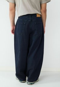 Luźne, ciemnoniebieskie dżinsy o wysokim stanie, z delikatnymi tylnymi kieszeniami i taninową skórzaną metką na pasku.