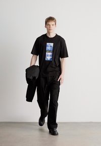 Μαύρη μπλούζα με γραφικό τύπωμα σε μπλε αποχρώσεις, συνδυασμένη με μαύρο cargo παντελόνι. Το μοντέλο κρατάει μια σκούρα ζακέτα και φοράει μαύρα παπούτσια.