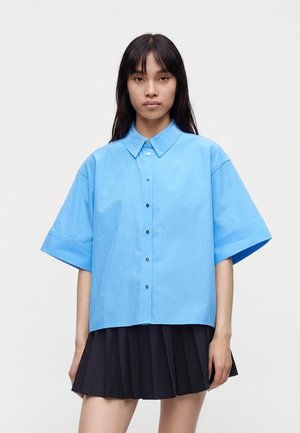 Jeune femme aux longs cheveux foncés portant une chemise ample bleu clair à manches courtes et boutonnée, ainsi qu'une jupe plissée noire sur fond blanc.