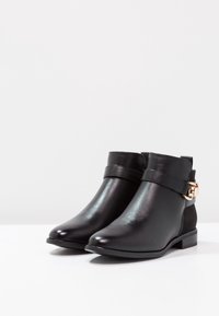 Par de botas negras hasta el tobillo con tacones bajos y una correa decorativa con una hebilla dorada sobre una superficie blanca.