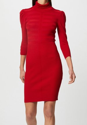 Femme portant une robe en maille rouge ajustée avec col roulé, manches trois-quarts et texture côtelée, se tenant devant un fond uni.