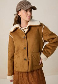 Bruine faux suede jas met een crèmekleurige, bont gevoerde kraag, voor knopen en twee zijzakken. Beschikt over een gestructureerde buitenkant en een zachte binnenkant.