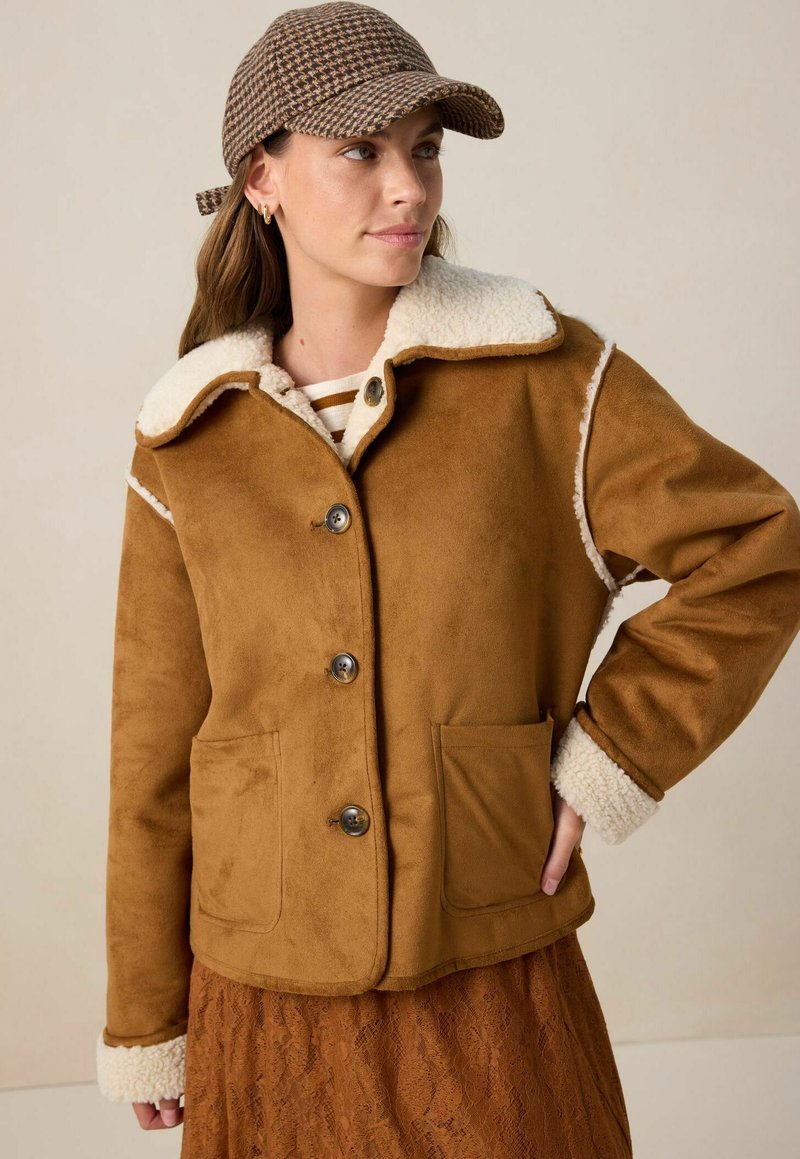 Bruine faux suede jas met een crèmekleurige, bont gevoerde kraag, voor knopen en twee zijzakken. Beschikt over een gestructureerde buitenkant en een zachte binnenkant.