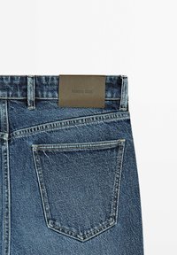 Denim jeans featuring a medium blue wash, five-pocket design, and a brown leather logo patch at the waistband.