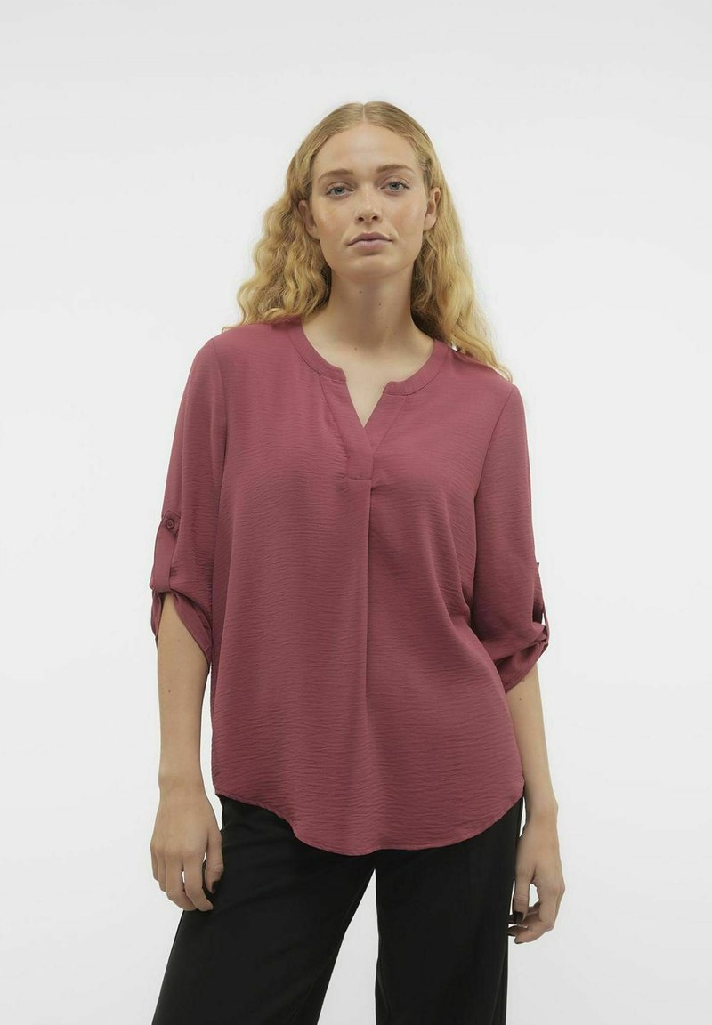 Top svasato a maniche corte in un profondo rosa con un tessuto testurizzato, scollatura a V e maniche arrotolate con bottone. Abbinato a pantaloni scuri.