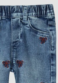 Pantalon en denim avec une ceinture élastique, ornée de nœuds rouges brodés à l'avant et denim bleu texturé avec une finition délavée.