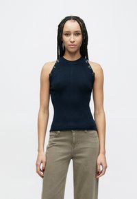 Top azul-marinho em malha canelada sem mangas com gola alta e detalhes de botões nas alças dos ombros, combinado com calças cáqui claras.