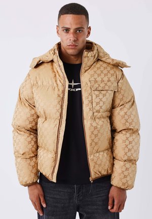 Young man wearing a tan patterned puffer jacket over a black shirt with logo, standing against a plain light background.