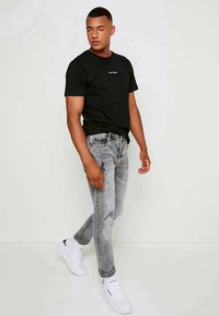 Jovem a caminhar, vestindo uma t-shirt preta, jeans cinza desgastados, meias brancas e ténis brancos, em frente a um fundo claro e simples.