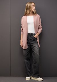 Ljusrosa kofta med ribbad kant, bärs öppen över en vit t-shirt, i kombination med mörkgrå jeans och krämfärgade sneakers. Slät struktur, avslappnad passform.