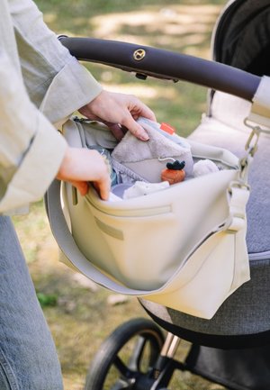 Beige-vaippalaukku, jossa on sileä pinta ja vetoketjutasku. Sisältää pehmeitä tavaroita ja pehmolelun. Kiinnitetty vaunujen kahvaan.