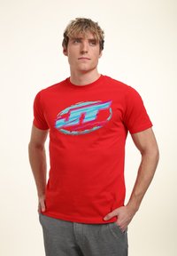 Červené bavlněné tričko s grafickým logem v modré a růžové barvě. Krátké rukávy, kulatý výstřih, slim fit design. Nosené s šedými kalhotami.