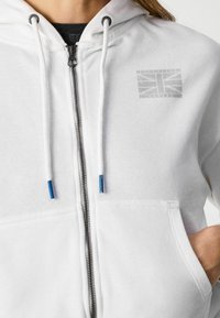 Felpa con cappuccio bianca con zip e tasca frontale, caratterizzata da un design grafico sottile, dettagli blu a contrasto sui lacci e una texture morbida.