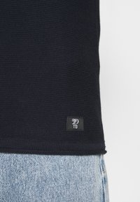 Pull en tricot bleu marine avec ourlet côtelé et petite étiquette noire avec texte blanc. Associé à un jean en denim bleu clair.