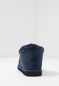 Marineblå ullfôr slippers med svart såle, sett fra baksiden, som viser merkeetiketten "Shepherd" på oversiden.
