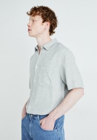 Chemise à manches courtes gris clair à boutons en mélange de lin avec une poche poitrine, portée avec un jean bleu en denim. Texture naturelle, coupe décontractée.